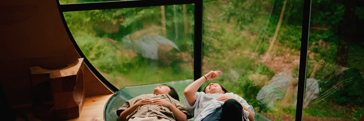 Two friends lying on a glass floor in a forest cabin, looking up at the trees
